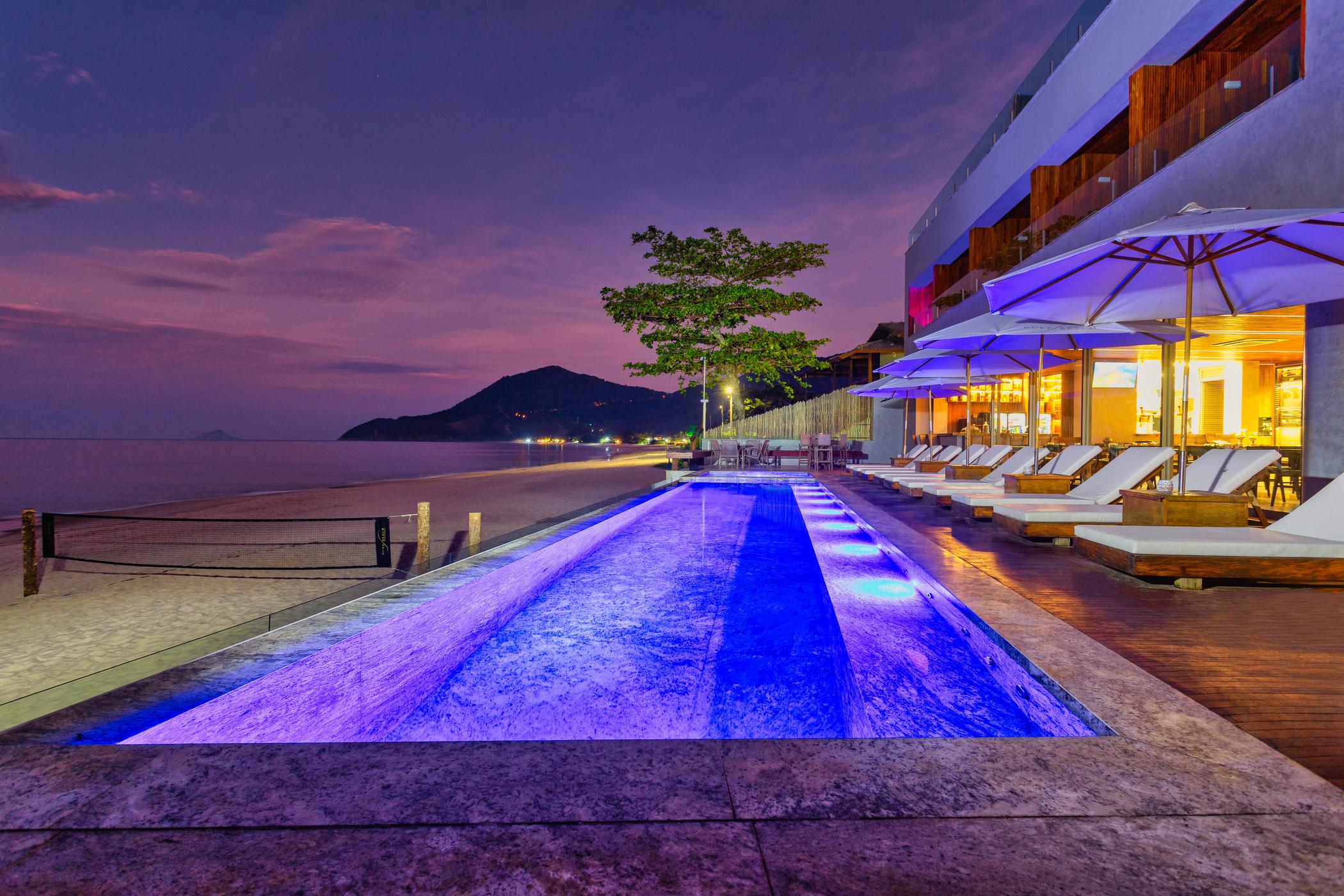 A piscina de borda infinita do Duke Beach ao anoitecer, a praia de Maresias à esquerda e o restaurante iluminado à direita.